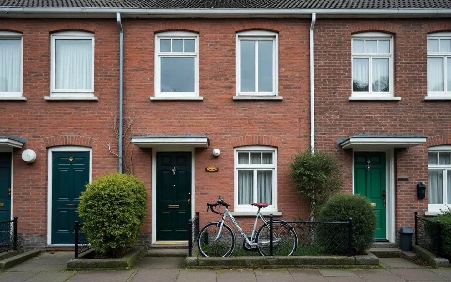Rij bakstenen rijtjeshuizen in zacht ochtendlicht. Bij één voordeur hangen de gordijnen dicht, staat een fiets tegen de gevel en steekt een brief uit de brievenbus.